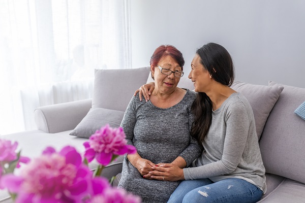 adult Asian daughter with her mature mother leaning on each other and being affectionate sitting on sofa at home.